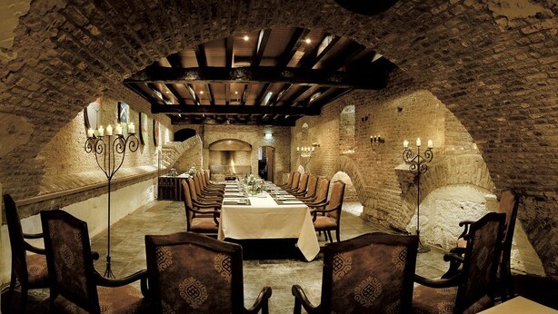 Una larga mesa de comedor con sillas, ubicada en una sala con paredes de piedra y techo abovedado.