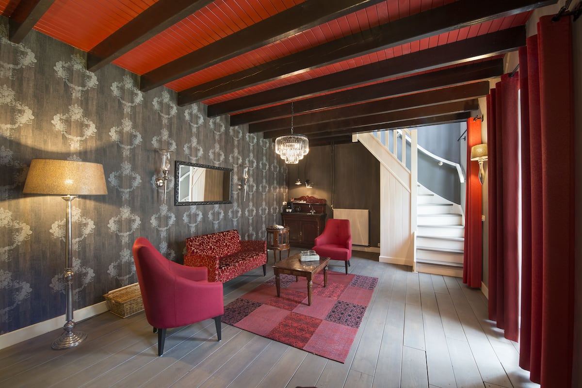 Une pièce avec un papier peint à motifs, un canapé rouge, un fauteuil rouge, une table basse en bois, un lampadaire et un lustre.