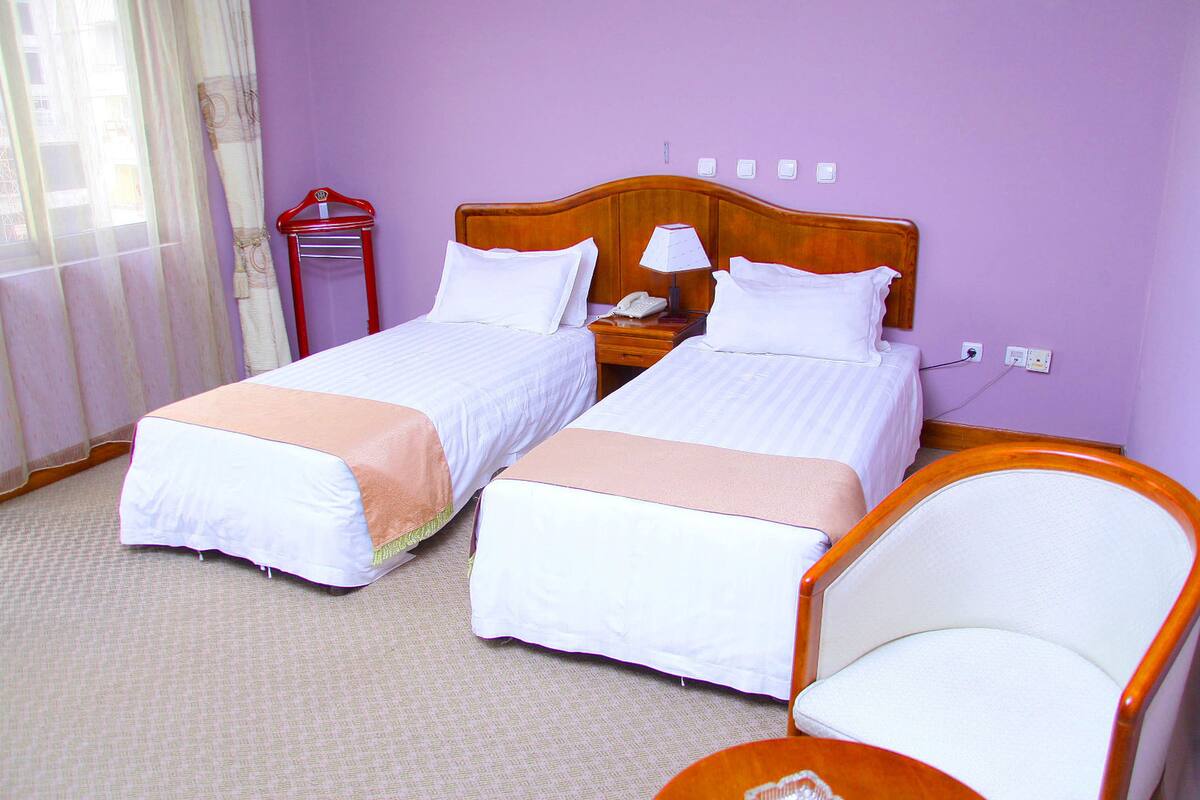 A hotel room with two beds, a nightstand with a lamp, a chair, and a window with curtains.