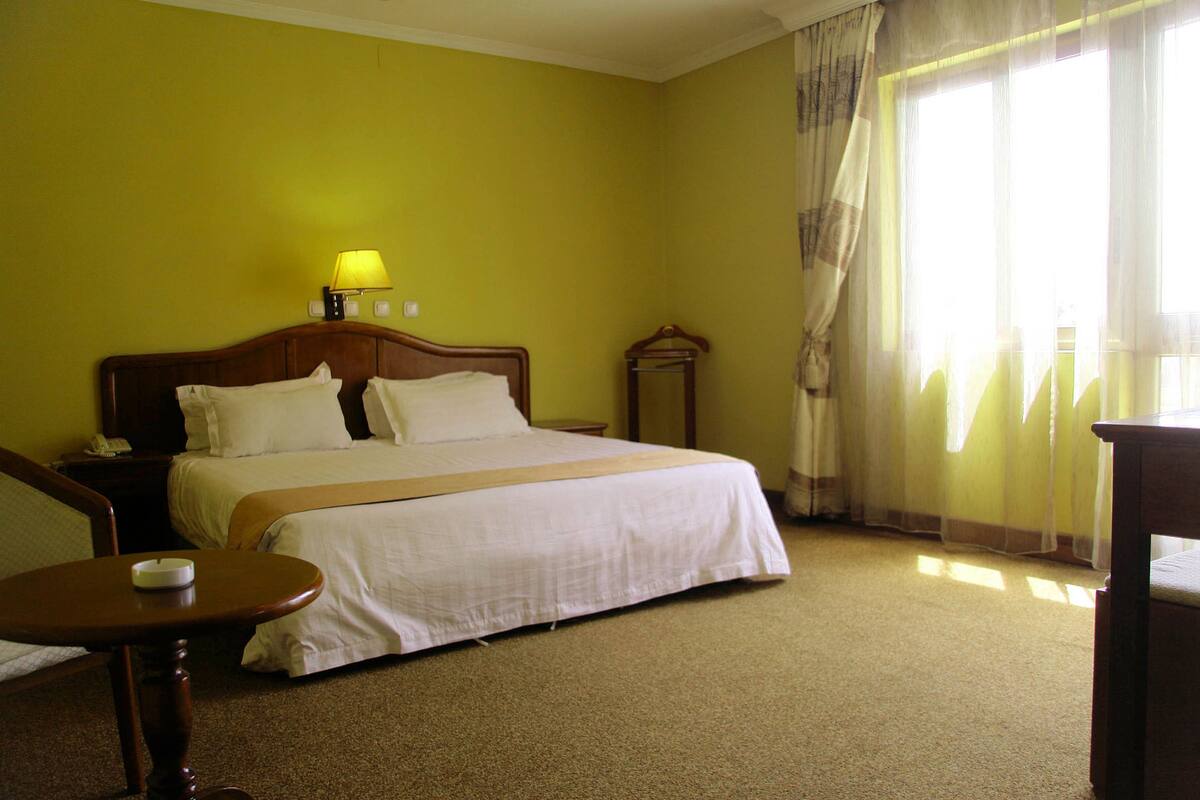 A hotel room with a large bed, a bedside table, a lamp, and a window with curtains.