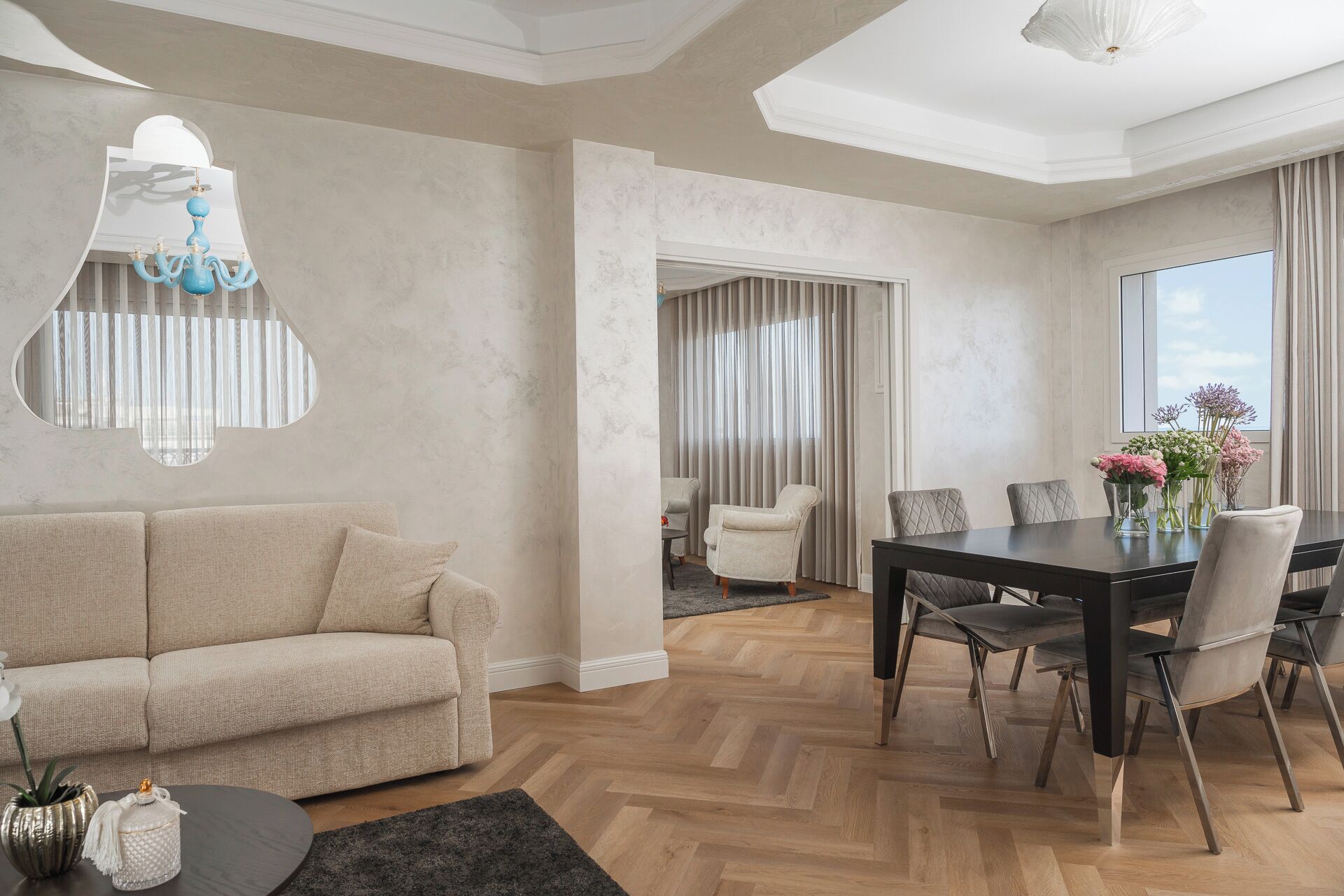 Ein modernes Wohnzimmer mit einer Couch, einem Esstisch und Stühlen. Eine Kristallleuchter, ein Fenster mit Vorhängen und eine Pflanze auf dem Tisch sind zu sehen.
