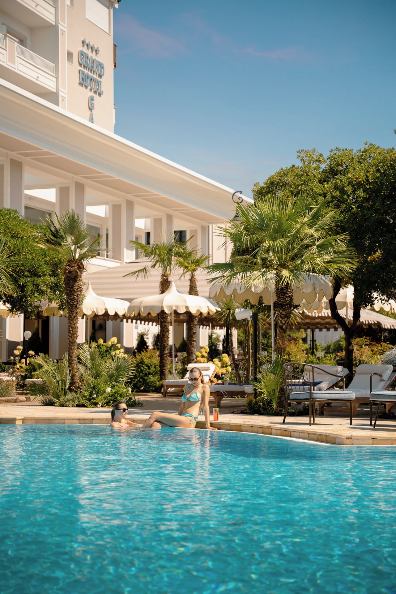 A swimming pool with lounge chairs and umbrellas, surrounded by palm trees and a hotel building.