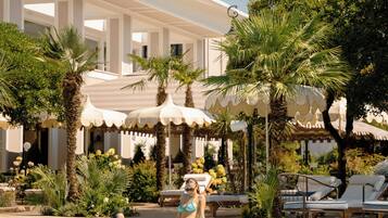 A swimming pool with lounge chairs and umbrellas, surrounded by palm trees and a hotel building.