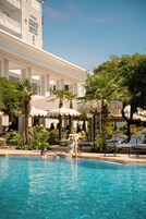 Una piscina con sillones de descanso y sombrillas, rodeada de palmeras y un edificio de hotel.