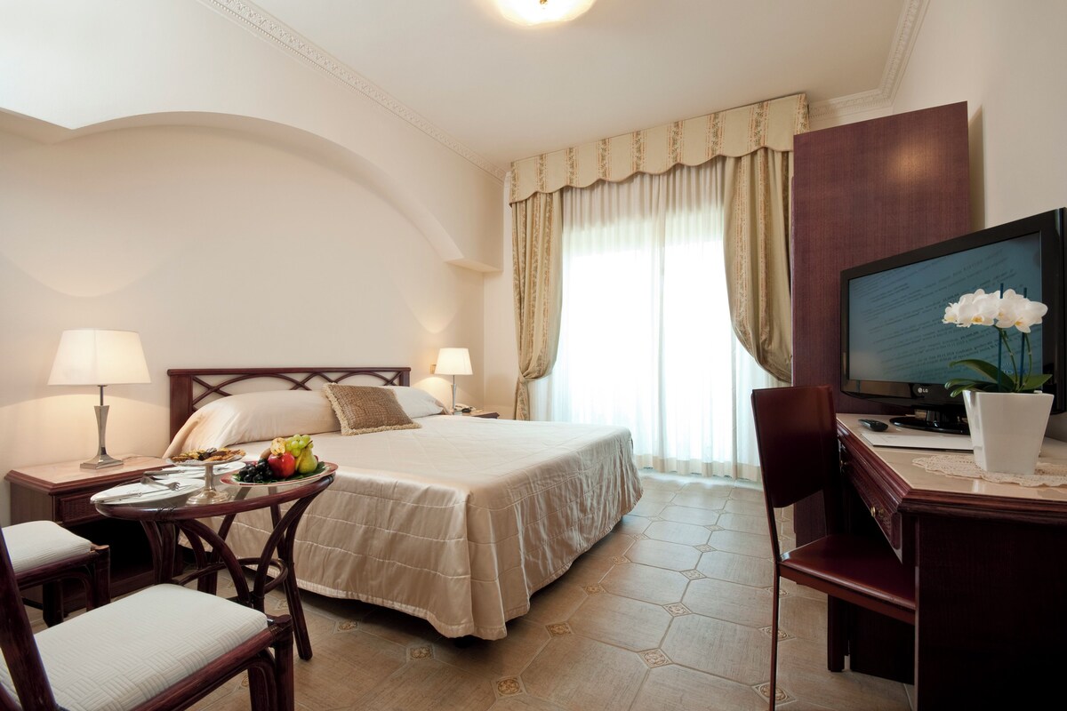 Habitación de hotel con cama, escritorio, silla, lámpara, televisión y una planta en maceta.