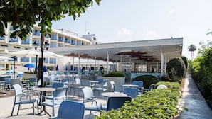 Un patio al aire libre con mesas y sillas, una zona de estar cubierta y un edificio con balcones al fondo.
