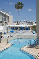 Una piscina con una palmera, sombrillas azules y un edificio con varios balcones al fondo.