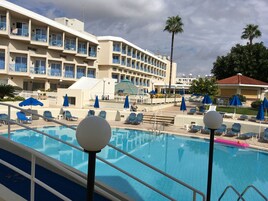 Zona de piscina de hotel con sillones de descanso y sombrillas, una palmera y un edificio con balcones.