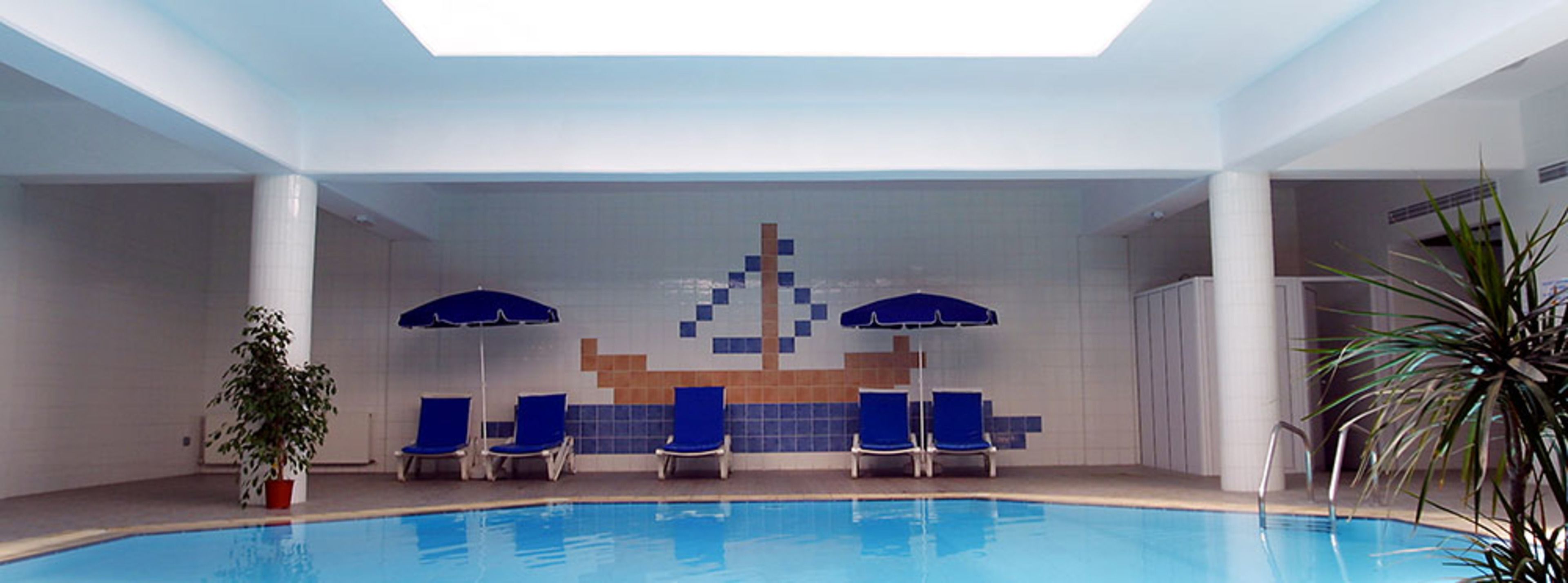 Une piscine intérieure avec des chaises et des parasols bleus, un mur carrelé orné d’un motif géométrique et une plante en pot.