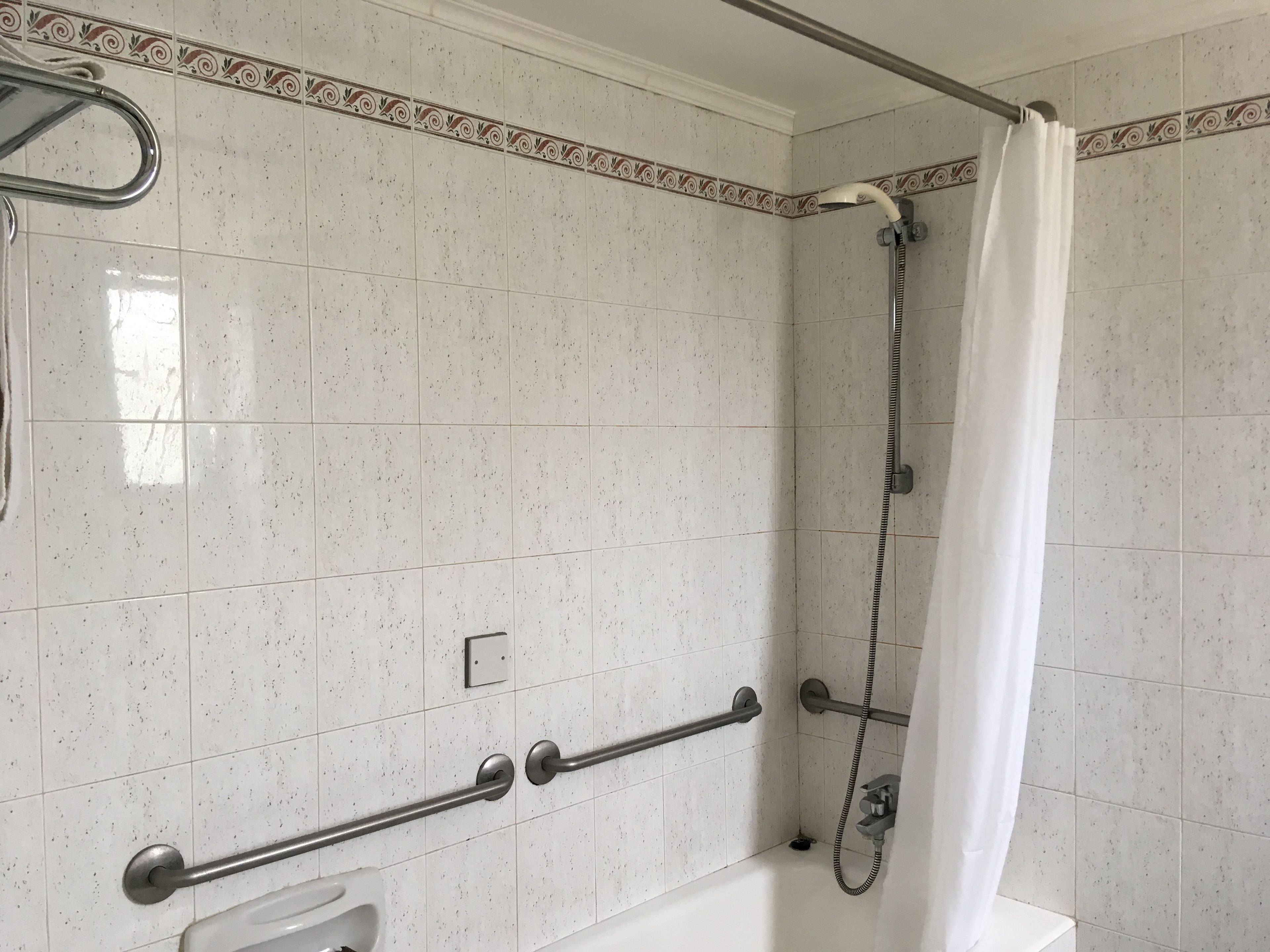 Un baño con baldosas blancas con motas, una ducha con cortina blanca y una barra de apoyo para seguridad.