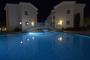 Una piscina con un puente que conecta dos edificios, rodeada de zonas bien iluminadas.