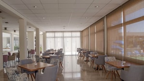 Un comedor amplio con varias mesas y sillas, ventanas grandes con cortinas translúcidas y un interior limpio y moderno.
