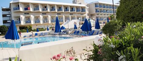 Um hotel com piscina, guarda-sóis azuis e um edifício de vários andares com varandas.