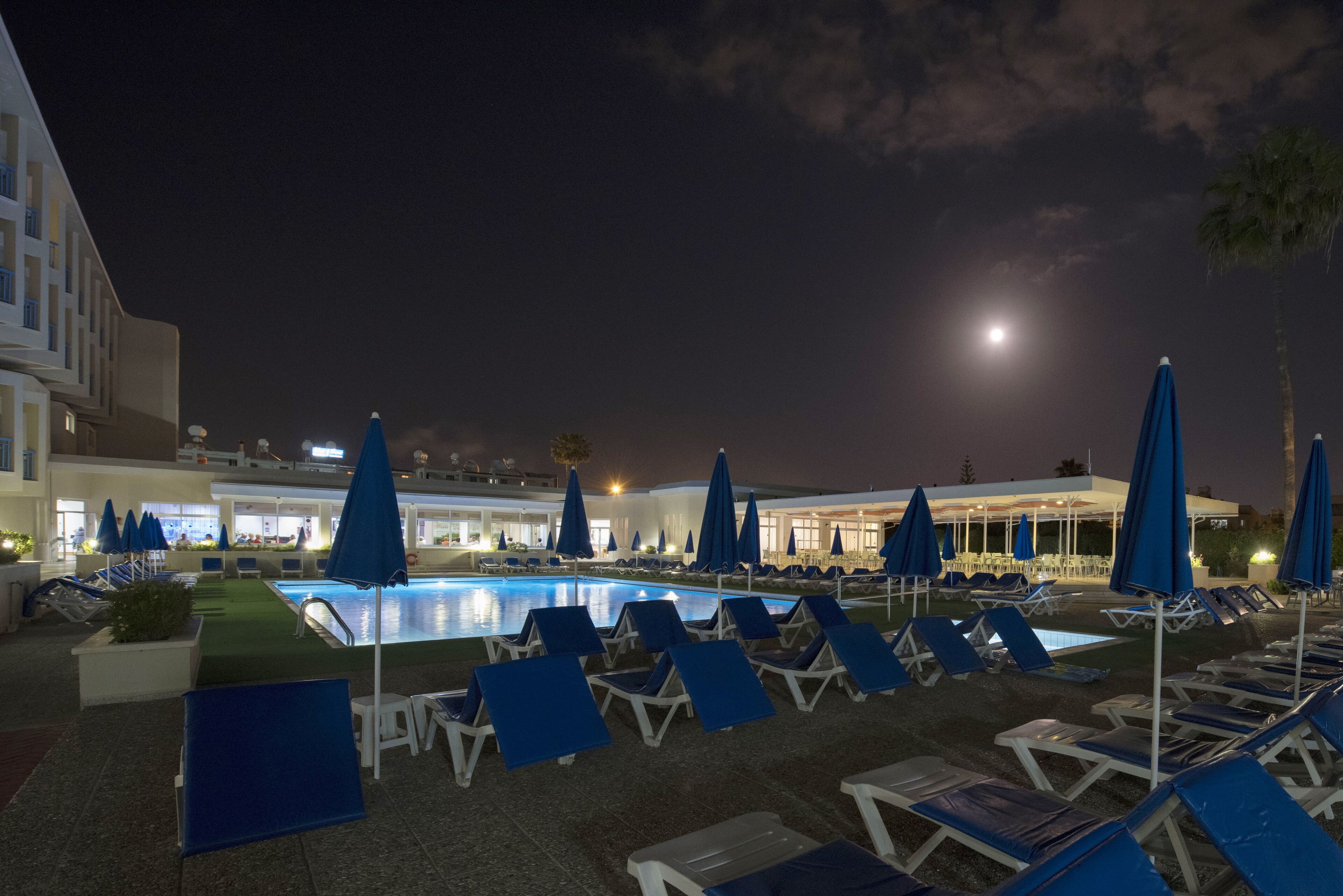 Uma área de piscina à noite, com espreguiçadeiras e guarda-sóis, piscina e um edifício iluminado ao fundo.