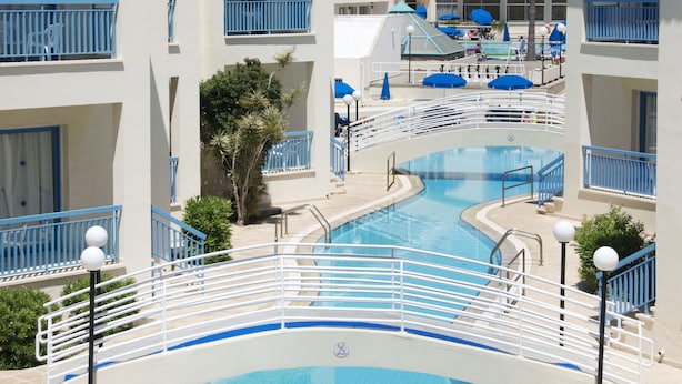Zona de piscina con un puente, rodeada de edificios con balcones y una palmera.