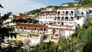 A white building with multiple balconies, a tiled roof, and surrounding greenery.