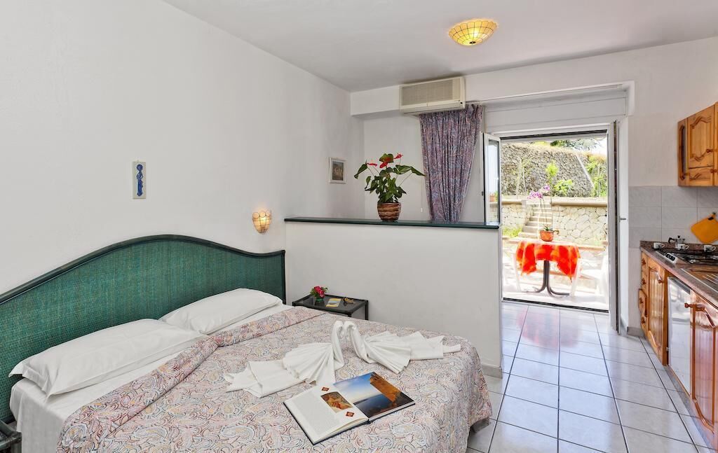 A bedroom with a bed, a bedside table, a potted plant, a window with curtains, and a view of the outside.