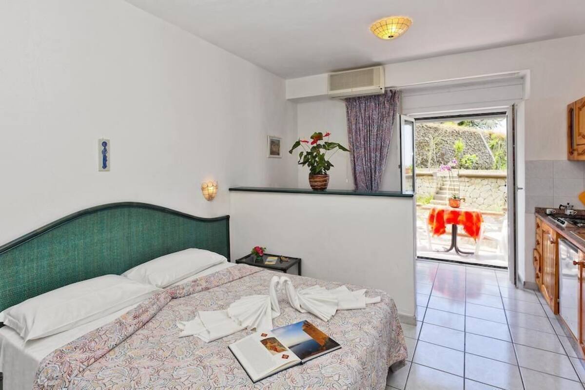 A bedroom with a bed, a bedside table, a potted plant, a window with curtains, and a view of the outside.