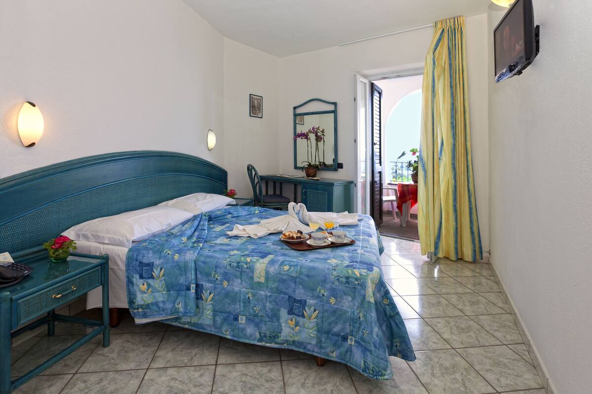 A hotel room with a bed, a desk, a chair, a TV, and a balcony with yellow curtains.