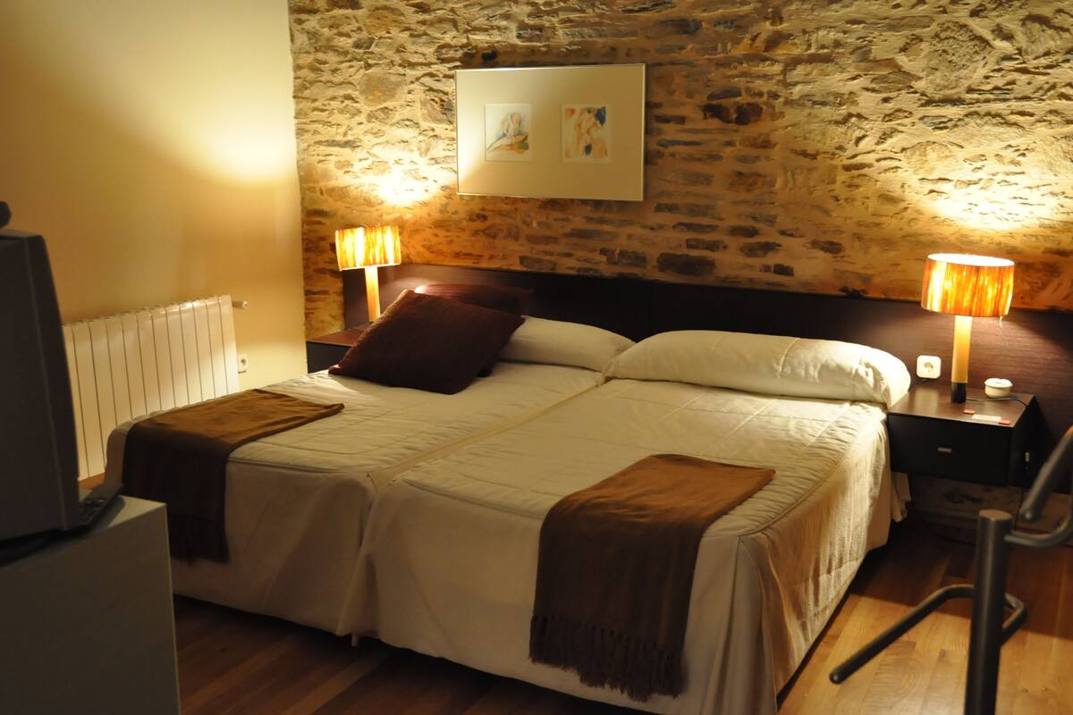 A bed with a headboard, two bedside tables with lamps, a radiator, and a stone wall with artwork.