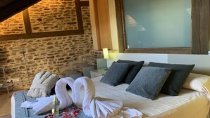 A cozy bedroom with a stone wall, a bed with white and grey pillows, a robe, and a glass of wine on the bed.