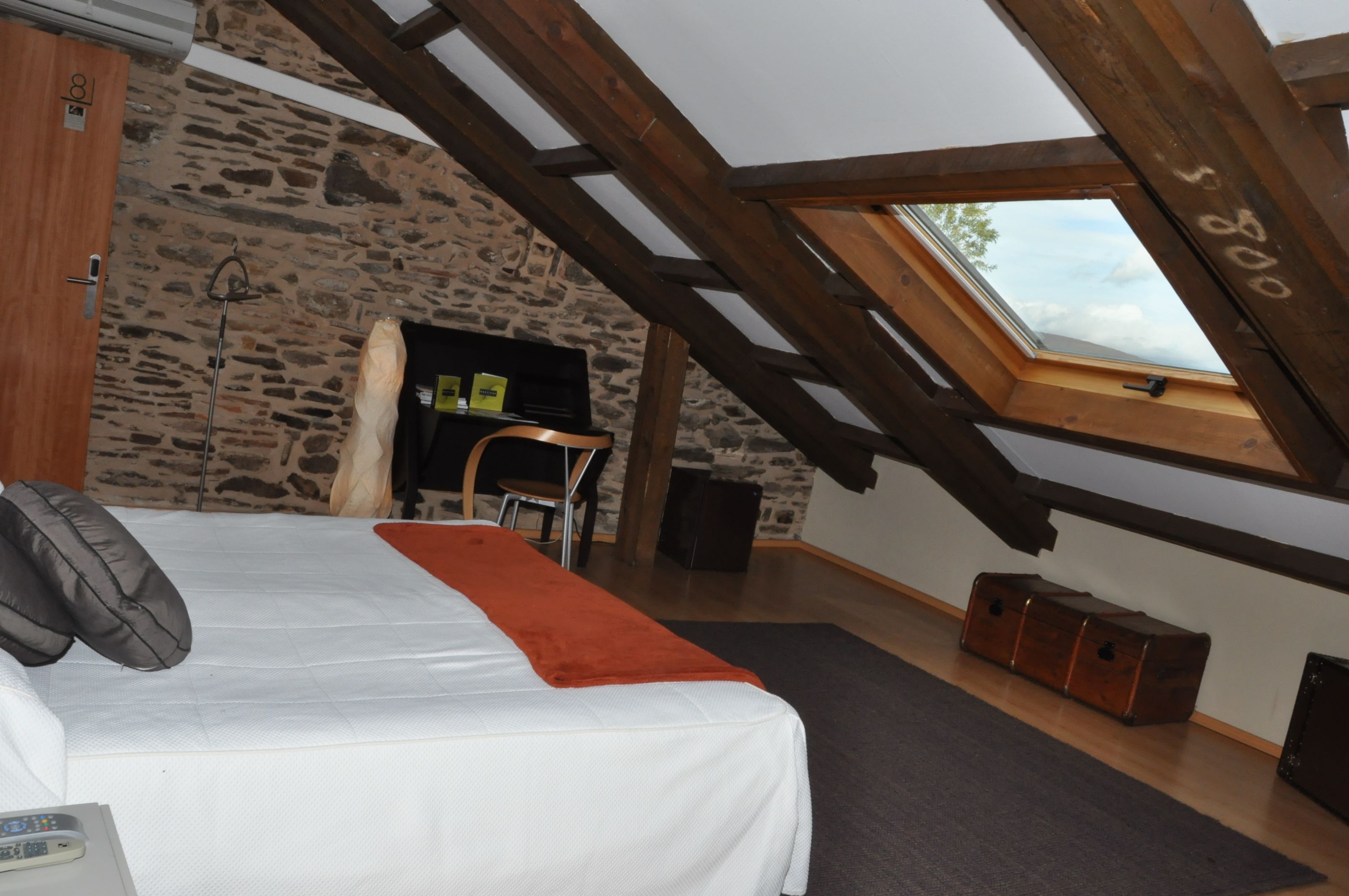 Un dormitorio con techo inclinado, pared de piedra, vigas de madera, una cama con almohadas, una mesita, una silla, una chimenea y una ventana con vistas.