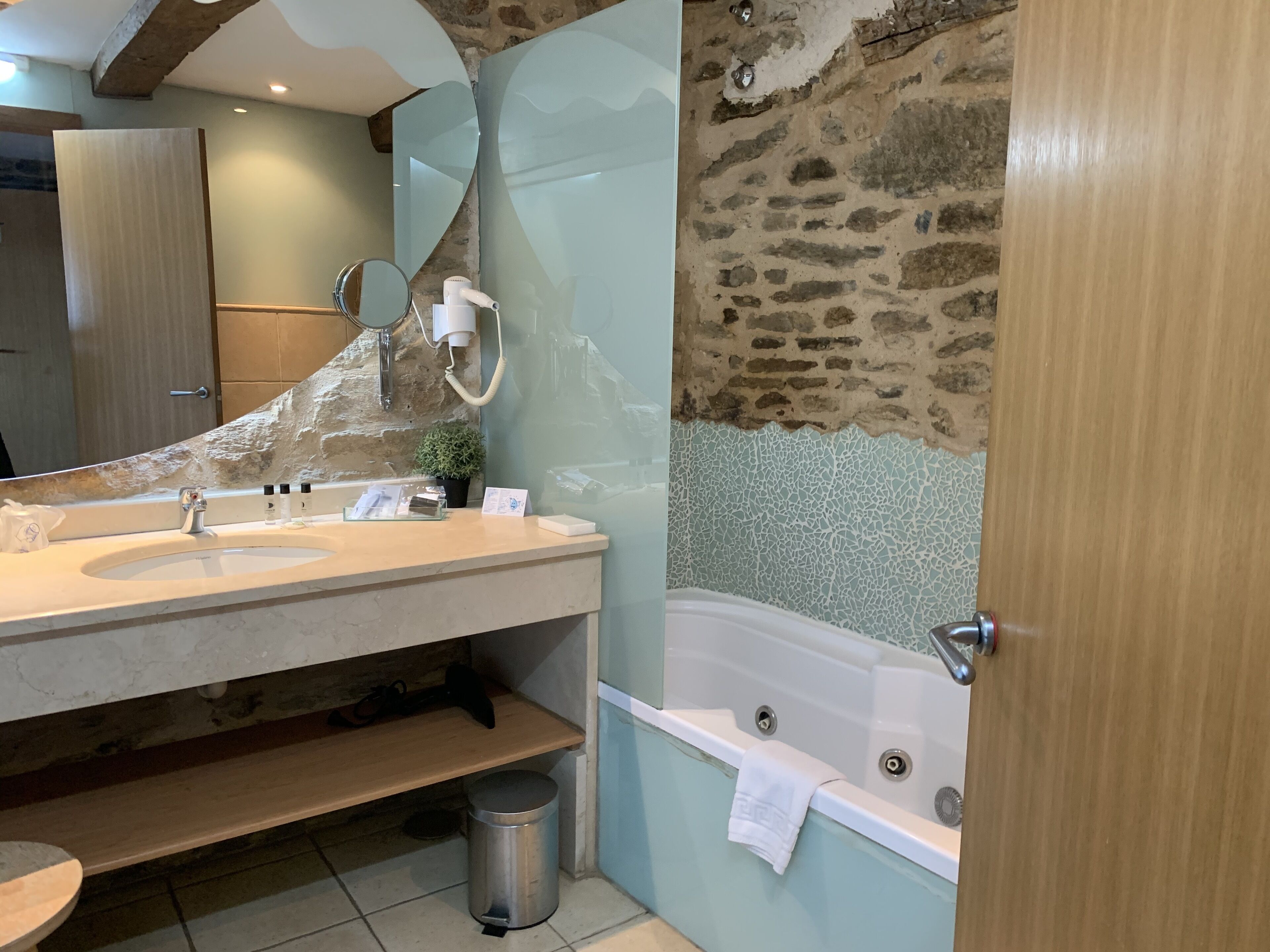 Un baño con pared de piedra, una bañera, un lavamanos con espejo y una pequeña planta en maceta.