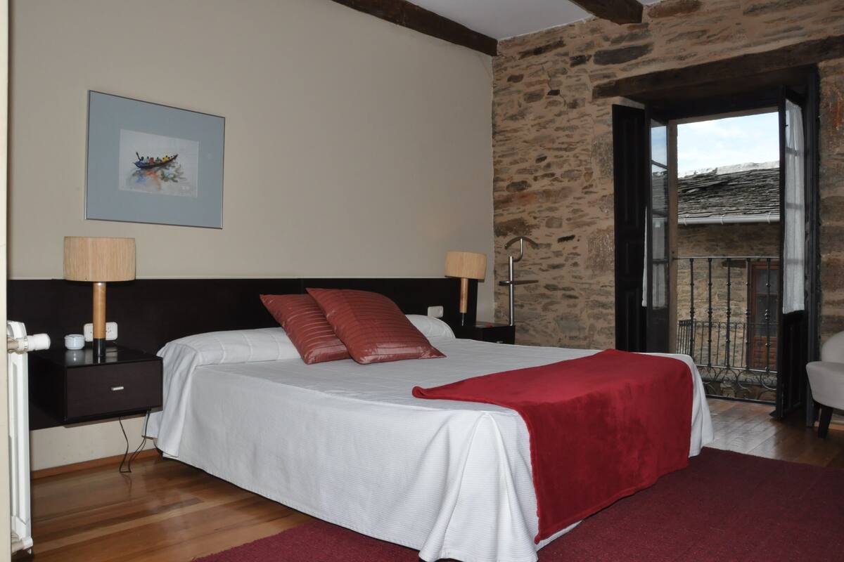A bedroom with a bed, a nightstand, a lamp, a picture on the wall, and a view of the outside.