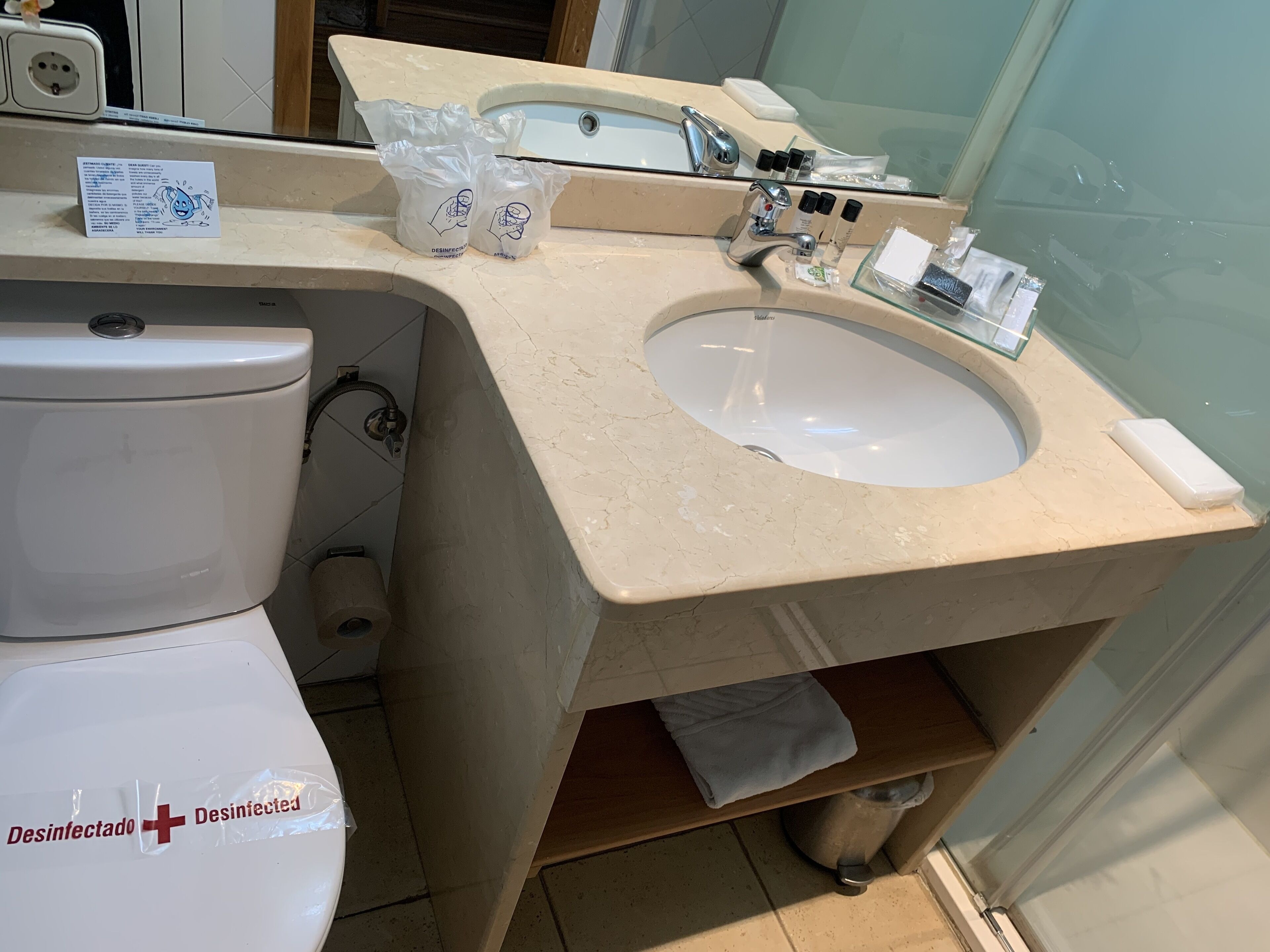 Un baño con inodoro, lavamanos y espejo. Hay bolsas desinfectantes y un cartel sobre el lavamanos.
