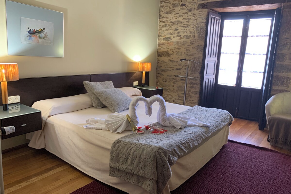 A neatly made bed with a swan towel arrangement, a framed picture on the wall, and a stone wall.