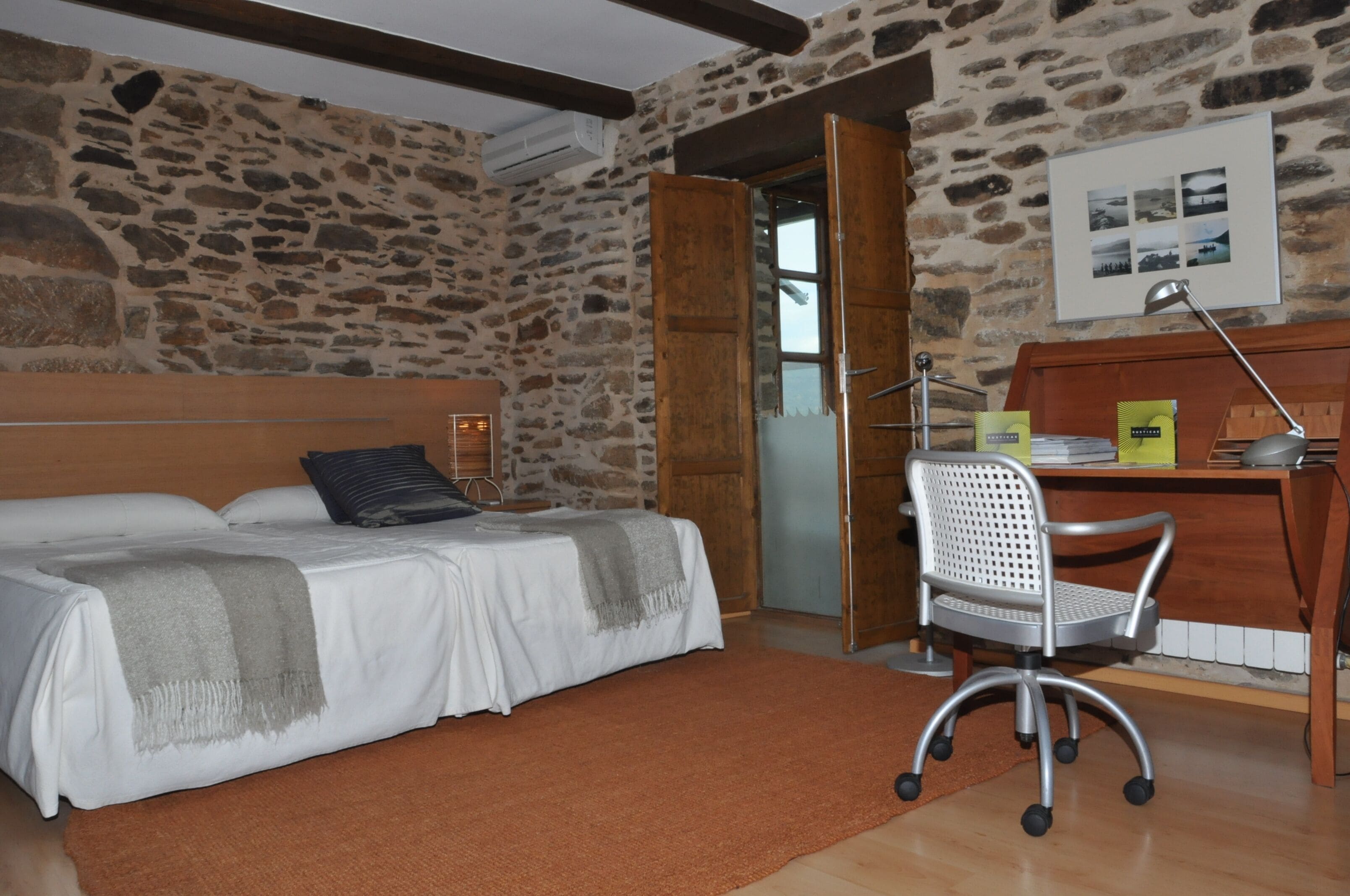 A bedroom with a stone wall, a bed with a wooden headboard, a desk with a chair, and a window.