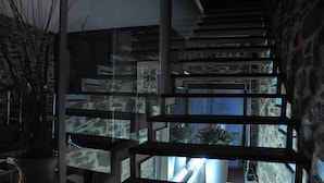 A staircase with stone walls and a glass railing, leading to a balcony with a view of the outside.