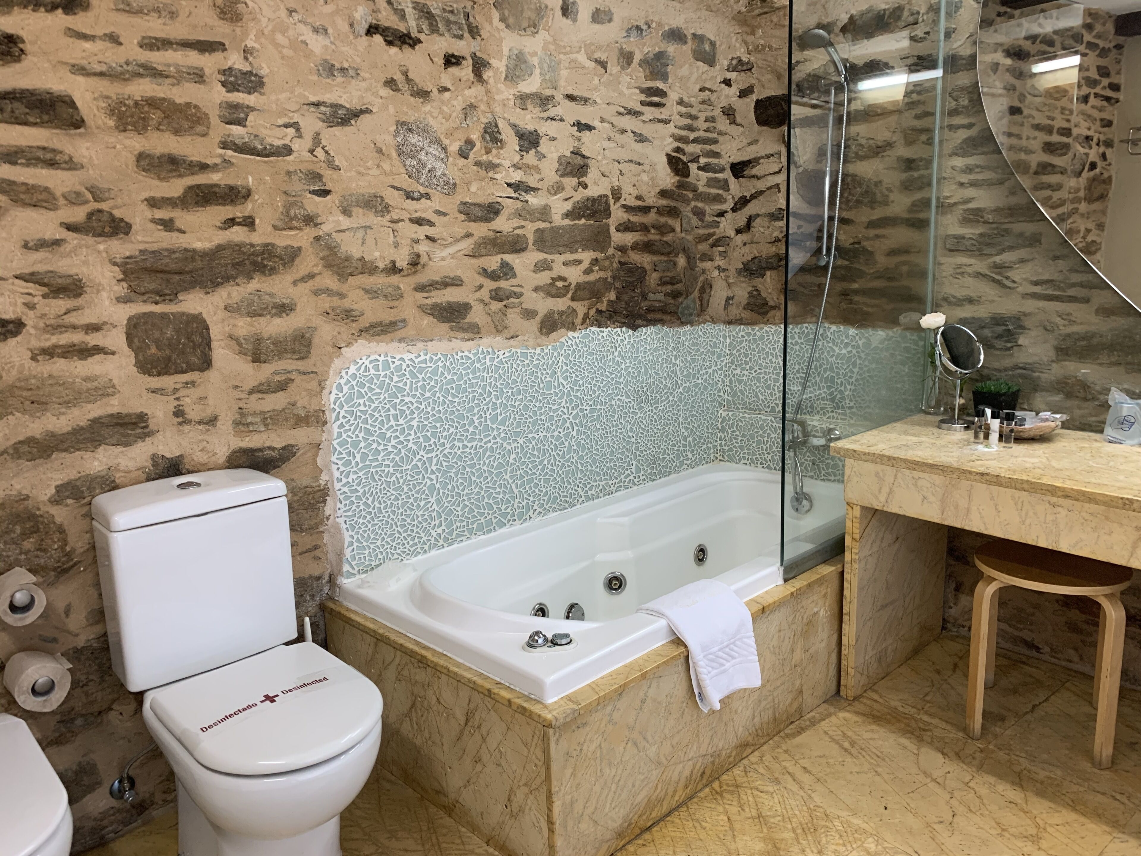 Un baño con pared de piedra, inodoro blanco, bañera y taburete de madera.
