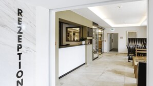 Un espace d’accueil avec un mur en marbre, un comptoir de réception, un coin salon avec une table et des chaises, et un miroir.