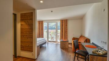 Una habitación moderna y bien iluminada con un revestimiento de pared de madera, una cama, un comedor con mesa y sillas, y vistas a un balcón con mobiliario exterior.