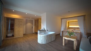 Une chambre d’hôtel moderne avec un plancher en bois, une baignoire ronde, un banc en bois, un lit avec des rideaux blancs et une petite table agrémentée de fleurs.