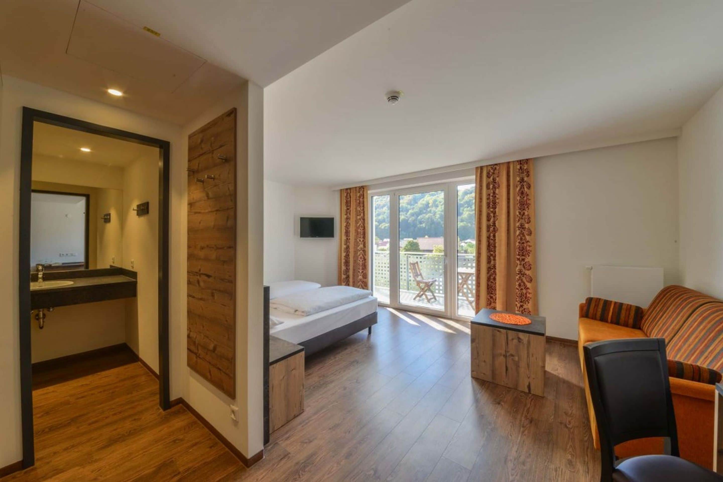A compact room with a bed, a small desk, a sofa, and a sliding glass door leading to an outdoor area.