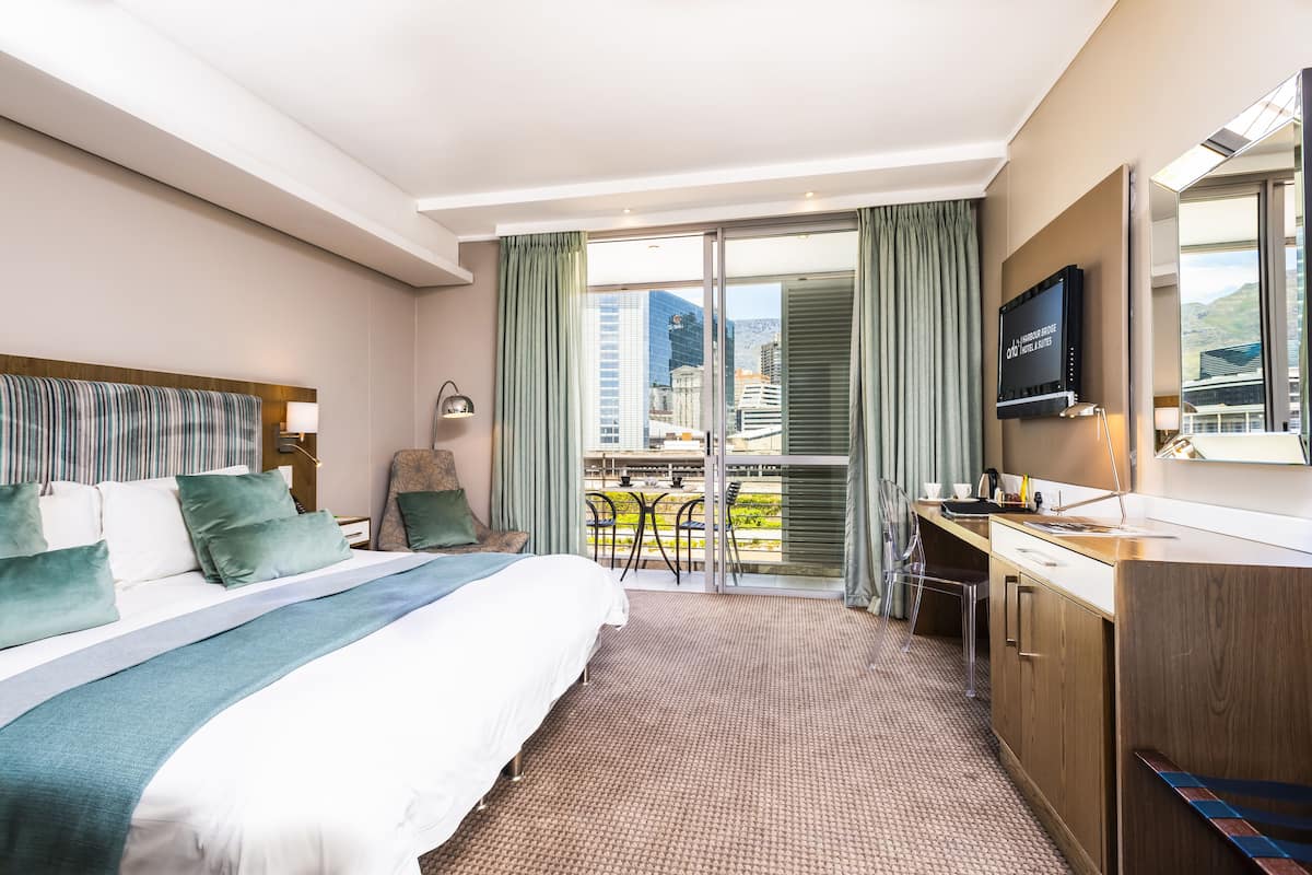 A hotel room with a large bed, a desk, a TV, and a balcony with a city view.