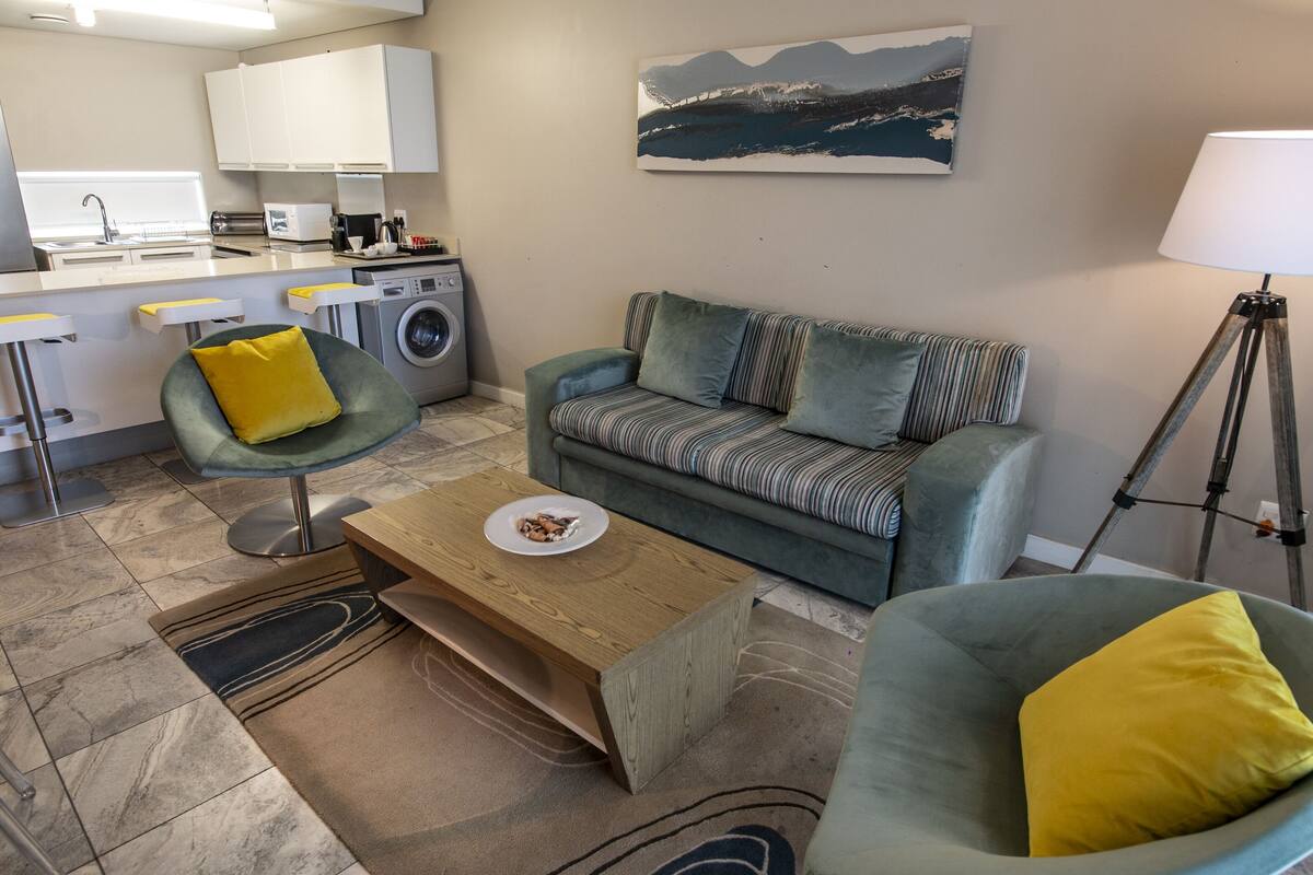 A modern living room with a sofa, armchairs, a coffee table, and a kitchen area with appliances.