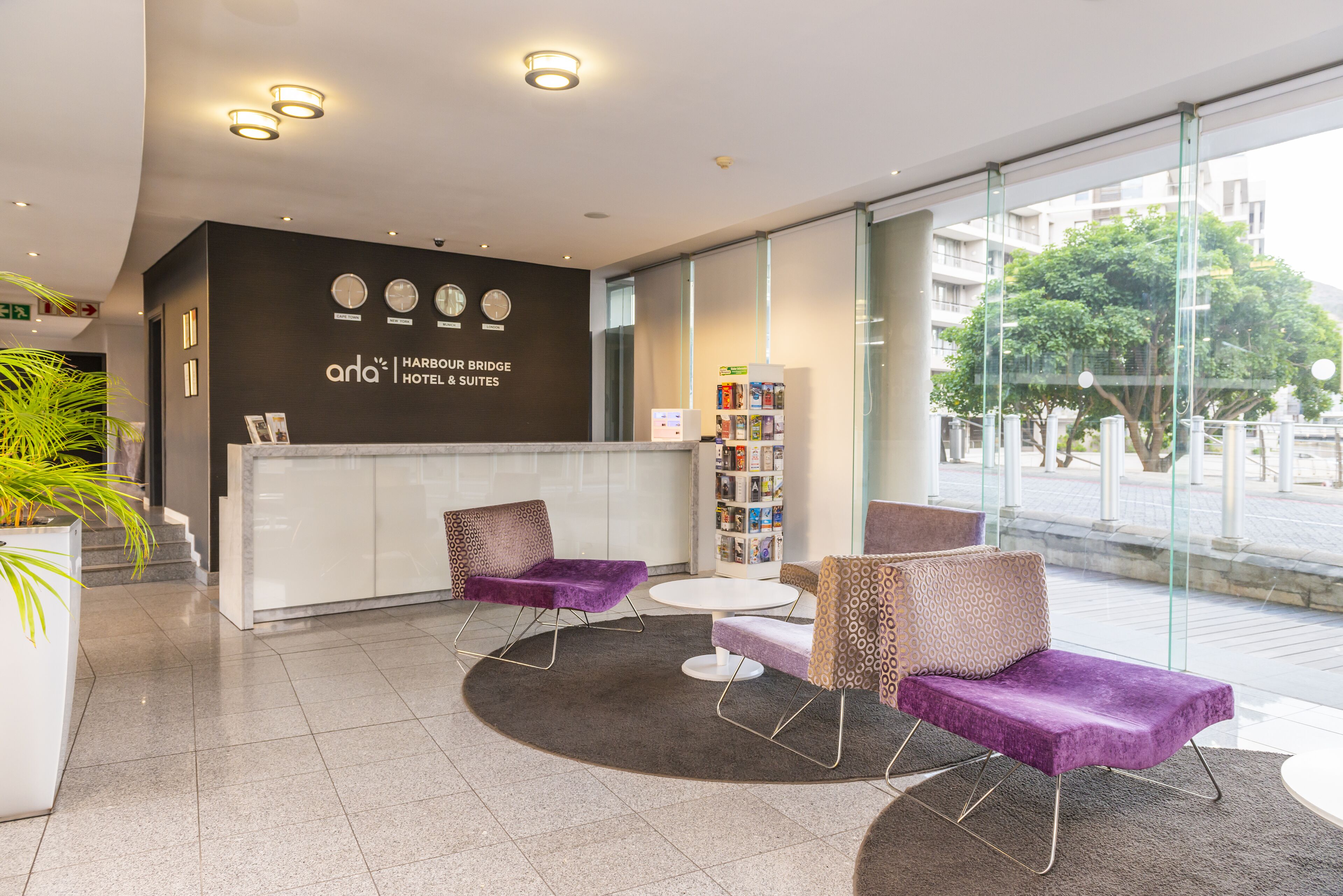 A modern hotel lobby with a reception desk, two purple armchairs, a small round table, and a bookshelf.