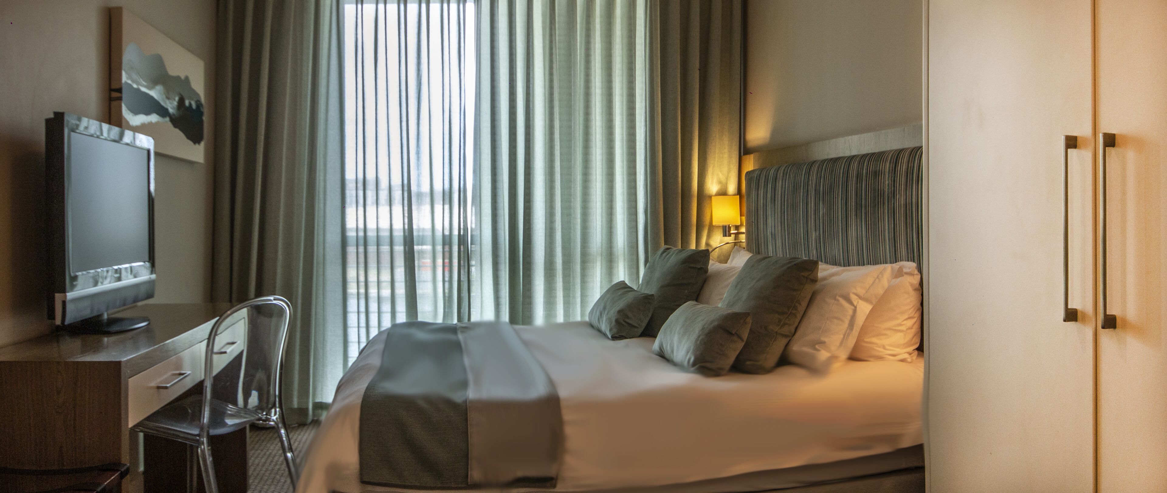 A hotel room with a bed, a desk, a chair, a TV, and a window with curtains.