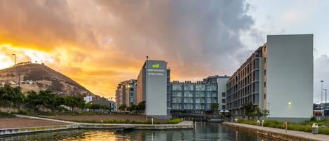 Un hotel moderno con un llamativo letrero de 'A&O', ubicado junto a una masa de agua tranquila, probablemente un canal, con un cielo de atardecer y un paisaje montañoso al fondo.