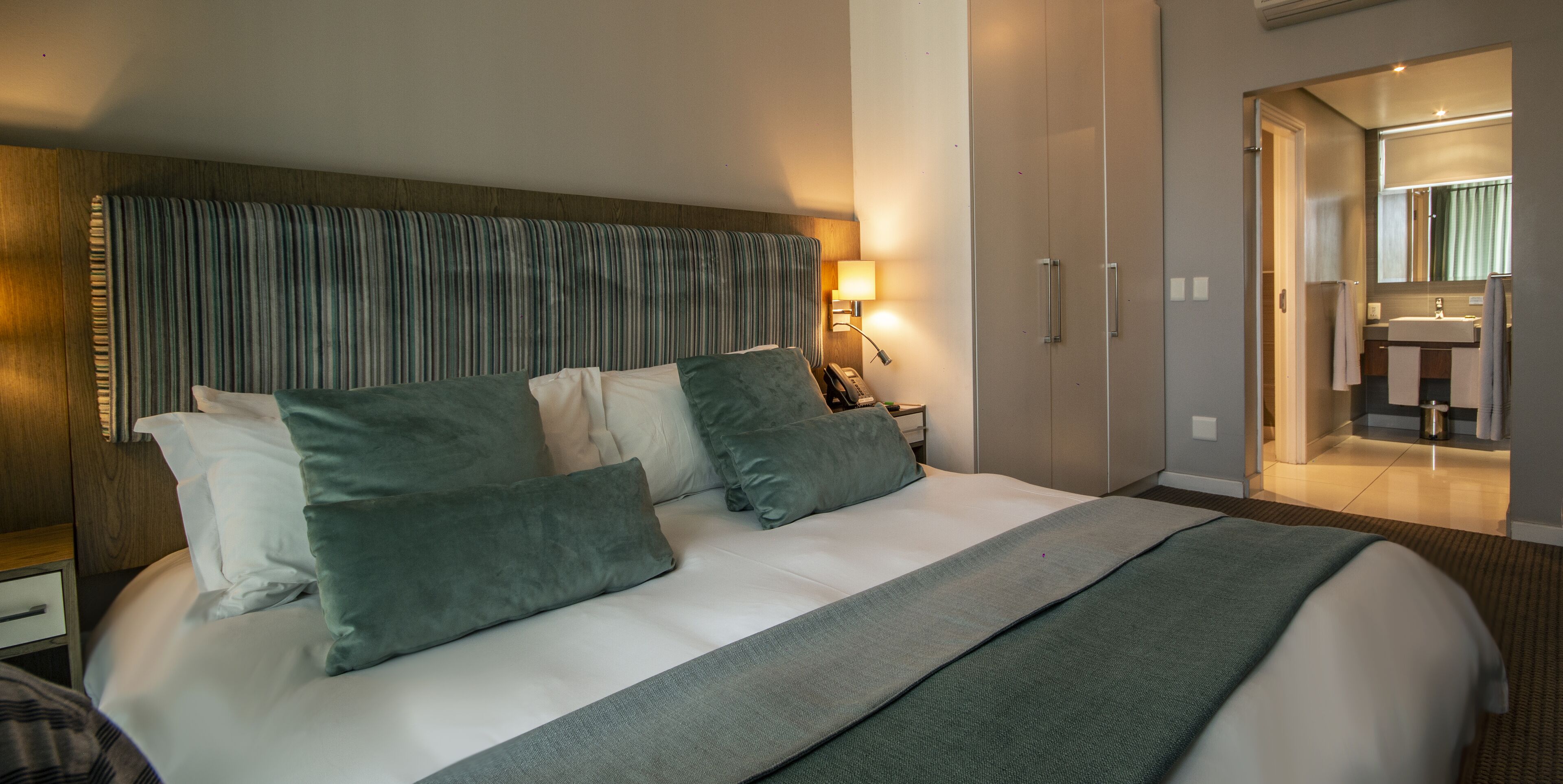 Quarto de hotel com uma cama grande, cabeceira, mesa de cabeceira com abajur e guarda-roupa.