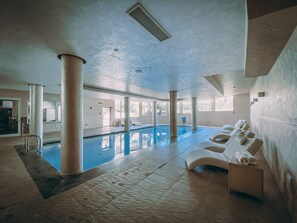 An indoor swimming pool with lounge chairs and a sunbed, surrounded by large windows and columns.
