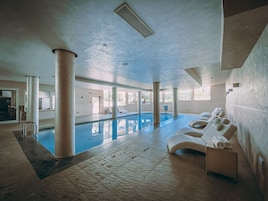 An indoor swimming pool with lounge chairs and a sunbed, surrounded by large windows and columns.