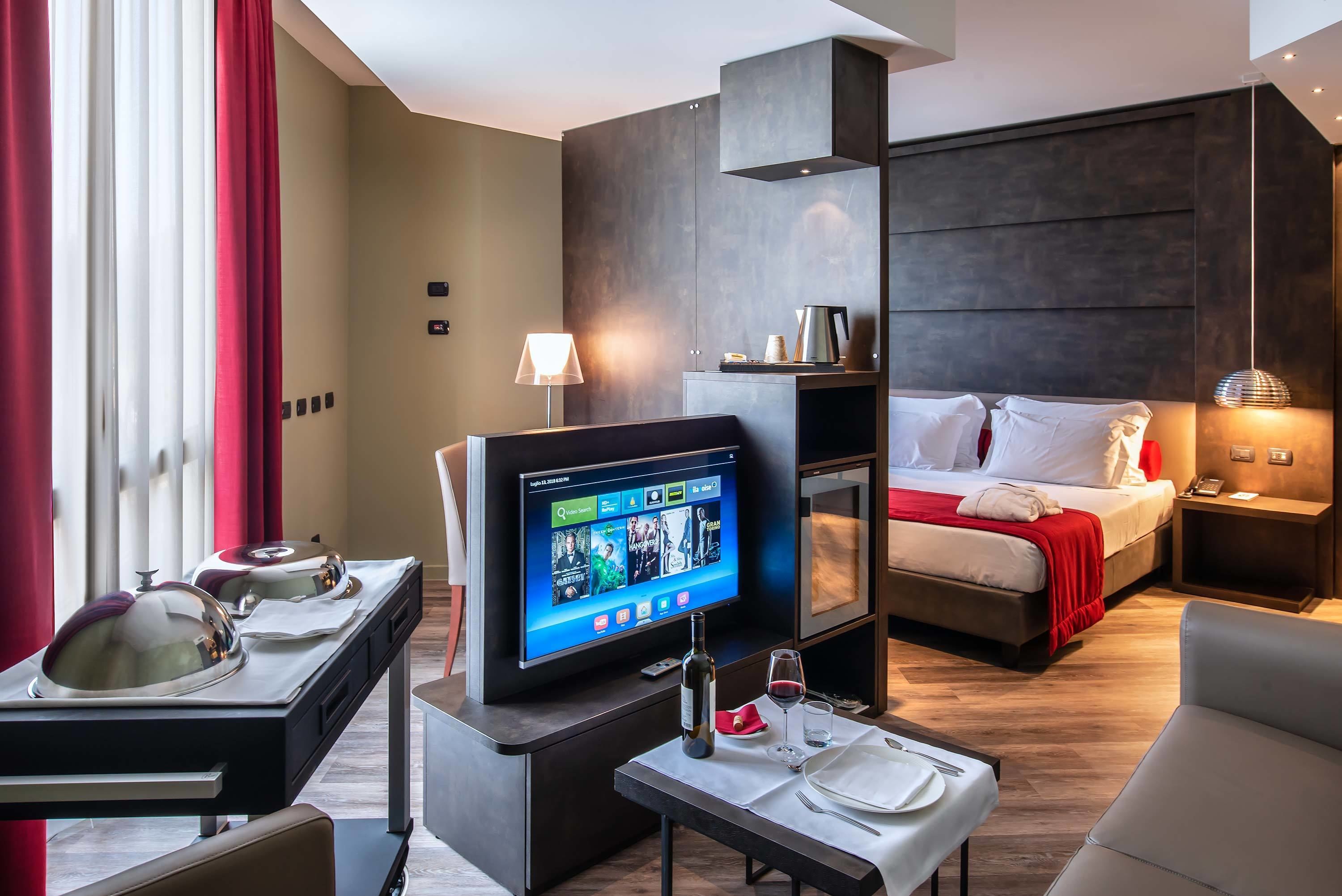 A hotel room with a bed, bedside table, TV, and a dining area with a table set for two.