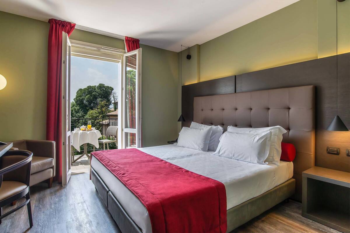 A hotel room with a bed, a desk, a chair, a nightstand, and a view of the outdoors.