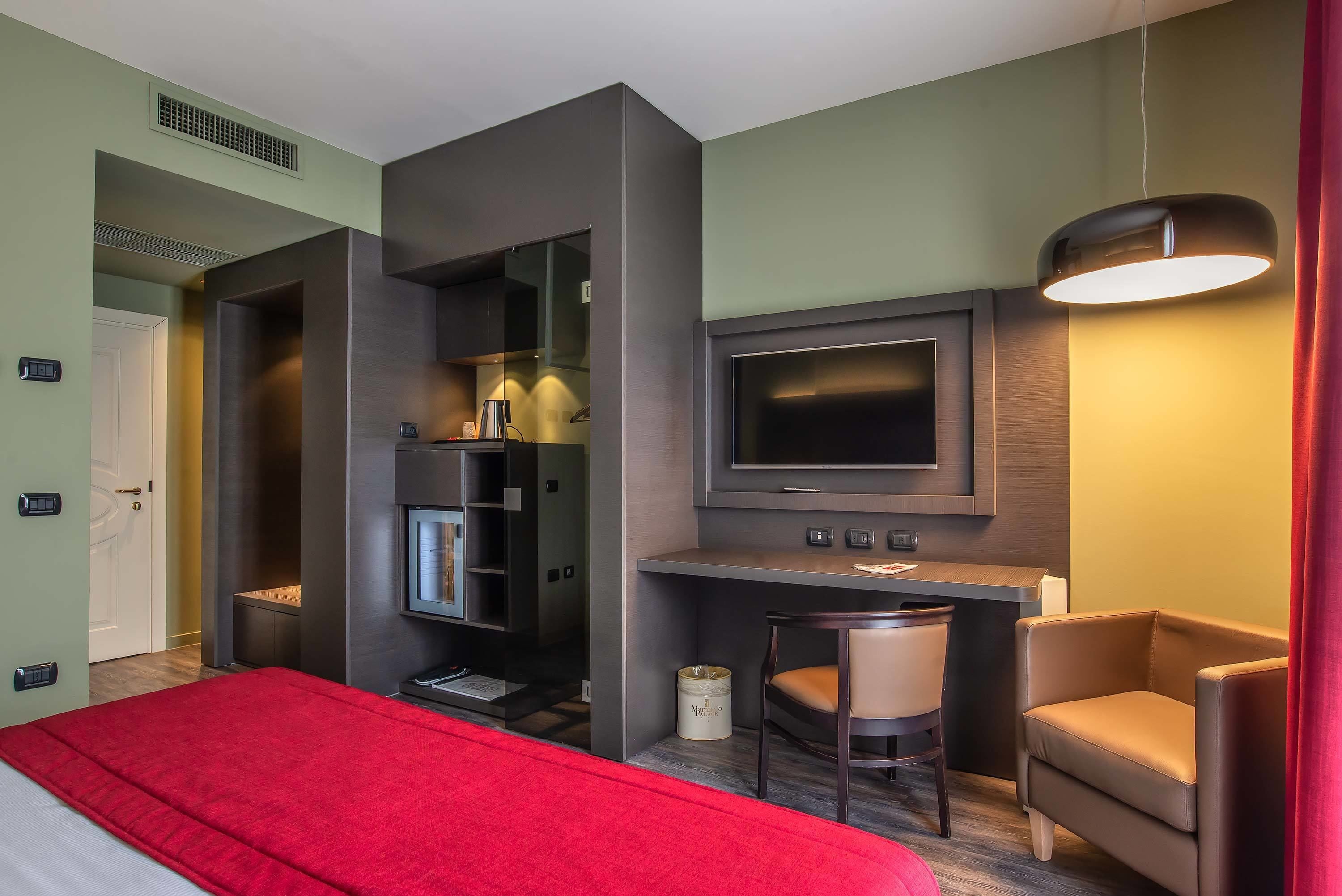 A modern hotel room with a bed, a desk with a chair, a television, and a red curtain.