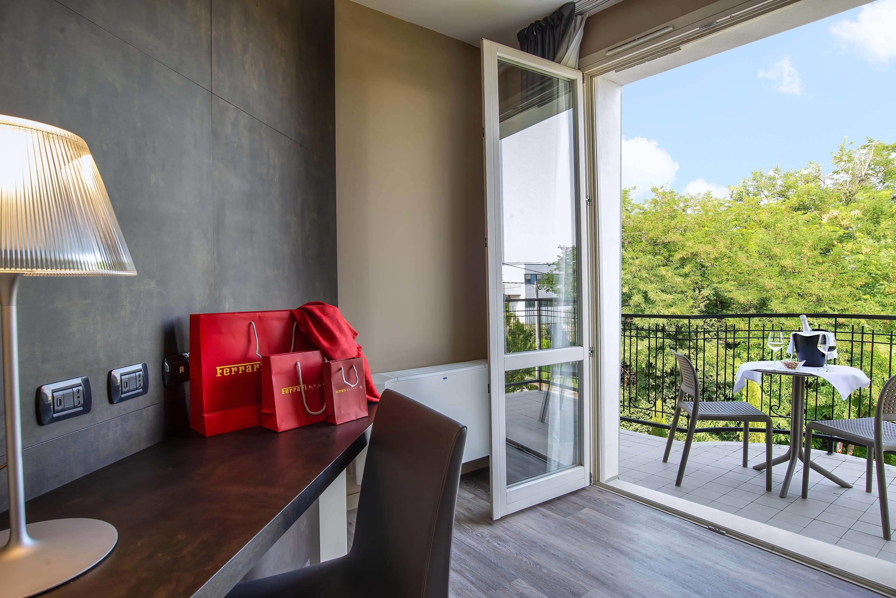 Habitación de hotel con un escritorio de madera oscura, una lámpara y dos bolsas rojas sobre la encimera. Se ve un balcón con una mesa y sillas a través de una puerta abierta.