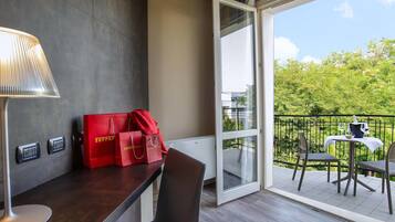 Habitación de hotel con un escritorio de madera oscura, una lámpara y dos bolsas rojas sobre la encimera. Se ve un balcón con una mesa y sillas a través de una puerta abierta.
